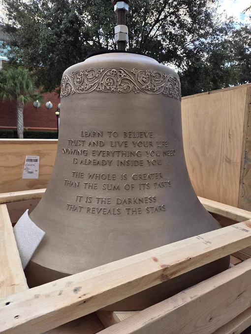 A large bell engraved with inspirational quotes, displayed in a wooden crate outdoors. 