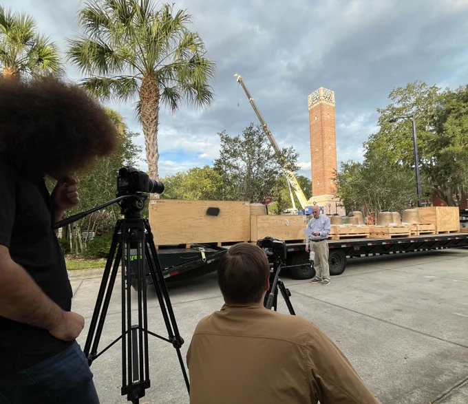 Two individuals filming Stetson's Chaplin,  who is speaking near a construction site with materials on a flatbed trailer, in front of a historical Hulley Tower.
