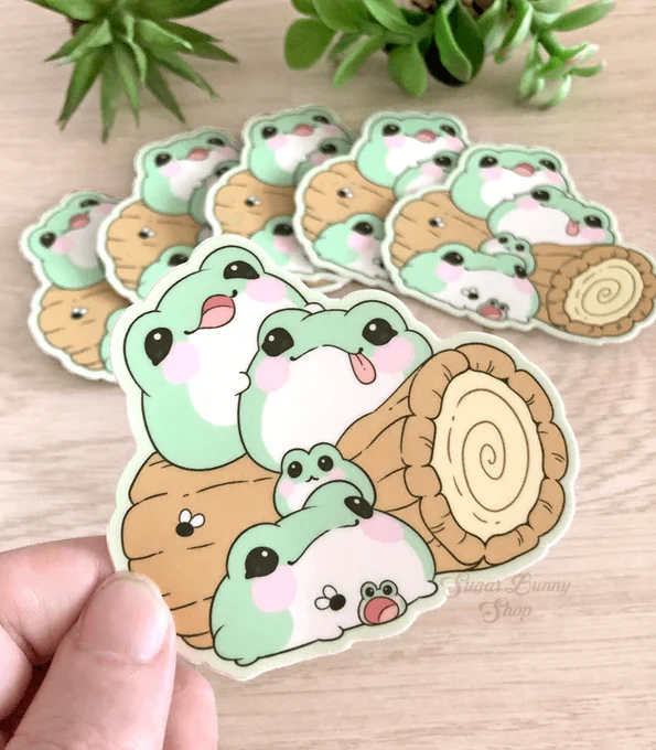 A hand holding a vinyl sticker featuring cute, fat blob-shaped frogs over a wood surface with plants in the background.