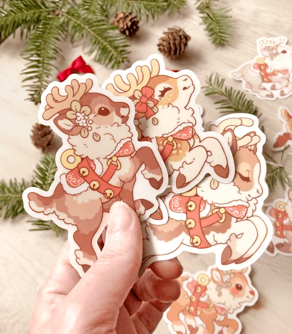 A photo of a hand holding three vinyl stickers featuring artwork of different designs for Santa's reindeer, held over a wood surface with pine branches.