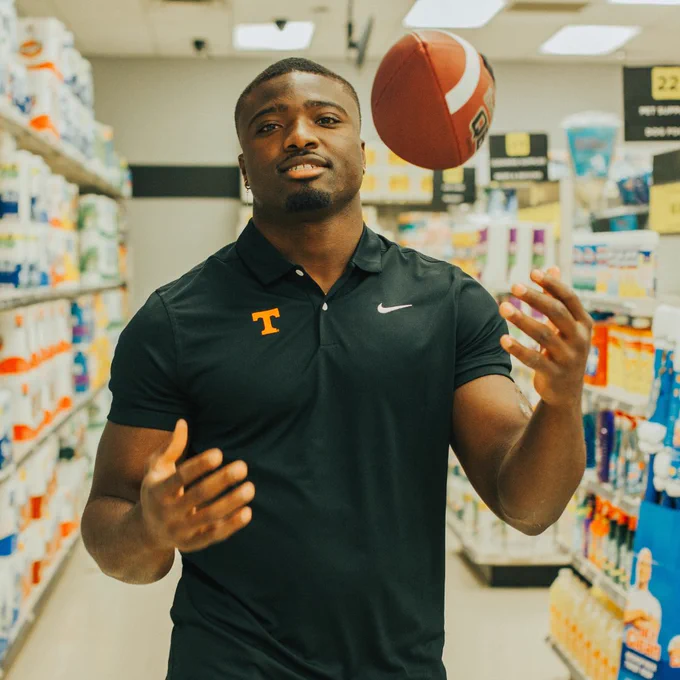Byron Young in a Tennessee polo shirt, with a Nike logo, stands in a store aisle, tossing a football in the air. Shelves are stocked with household items in the background.