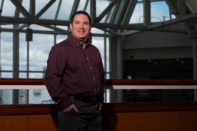 Dan Hogan in a dark red checkered shirt standing indoors with hands in pockets against a railing and large windows in the background.