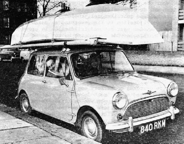An upturned boat on the roof rack of a classic Mini car. The driver is looking out of the sliding driver's window.
