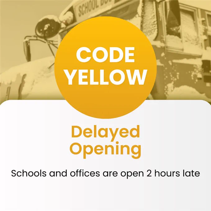 School bus covered in snow behind a notice announcing a Code Yellow delayed opening with schools and offices opening two hours late.