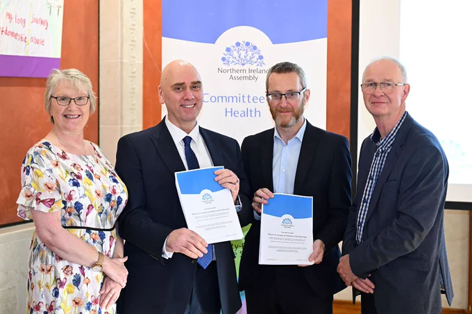 This photo shows 4 people, one woman in a floral dress and three men wearing dark suits, standing in a row. The two men in the centre are holding blue and white documents. Behind them is a pull-up banner with a blue and white logo of flax flowers at the top and the words 'Northern Ireland Assembly' in back text underneath. Underneath that are words in blue text which read 'Committee for Health'. 