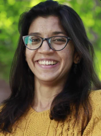 Head and shoulders photo of Neelima Sharma, having long dark hair, brown face, smiling, wearing dark-framed spectacles and yellow jumper against a green background