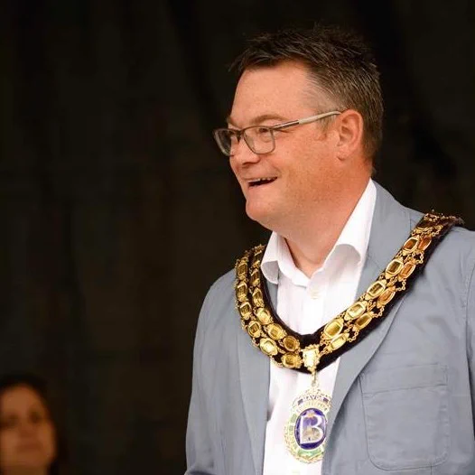 A man wearing glasses, a light grey jacket and ceremonial chains of office stands in a meeting. He is delivering a speech.