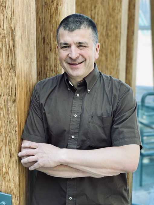 Person smiling at the camera, standing with arms crossed leaning on a wooden column. The individual is wearing a brown shirt.