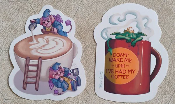 Photograph of two coffee themed stickers with little imps in or around the cups