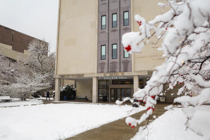College Hall with snow on the lawn and in the air. A student is next to the building walking to the Student Union.