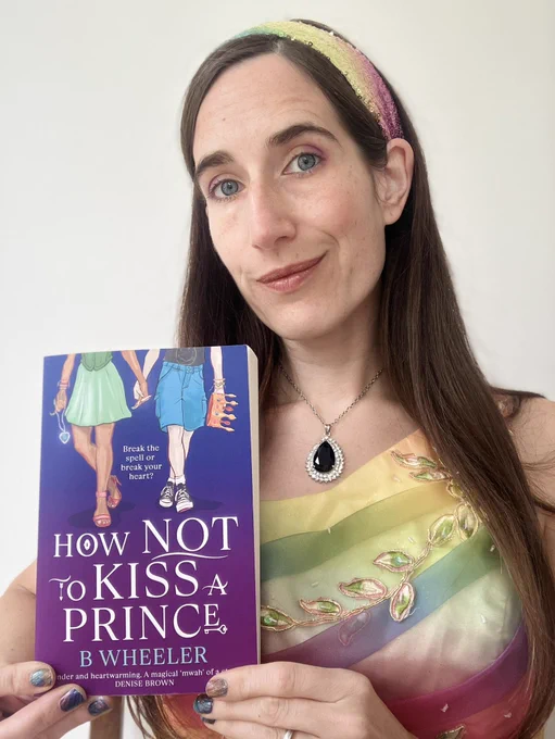 Author wearing rainbow dress holding paperback of How NOT TO Kiss A Prince. Book has purple background with images of two girls from the waist down holding hands above the title in white font.