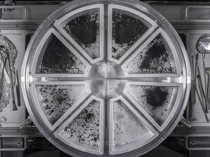 This image shows a circular metal container divided into six wedge-shaped sections, each filled with varying amounts of dark, fine-grained material and small rocks. The surface is highly organized, with scientific tools and utensils placed neatly to the left and right sides. This container holds samples collected from asteroid Bennu by NASA’s OSIRIS-REx mission, prepared for analysis in a sterile laboratory environment.