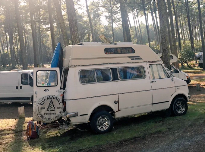 A white high-top Bedford CF camper in a wooded campsite. It has solar panels on the roof. 