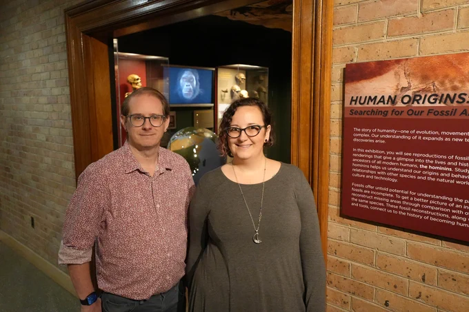 Two individuals smiling for the camera inside a museum exhibit titled "Human Origins: Searching for Our Fossil Ancestors."