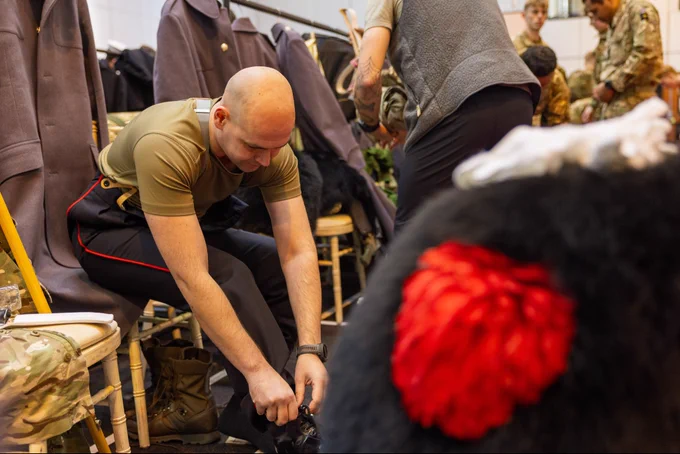 A seated soldier fastens his bootlace as he dresses in the changing rooms, surrounded by his uniform and red plumed bearskin cap