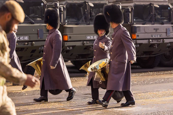 Army musicians wearing grey coats and bearskin caps arrive carrying brass instruments and walk to take up their place in rehearsals