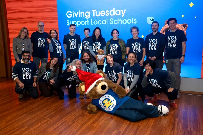 A group of people in volunteer shirts smile with a Build-A-Bear mascot wearing a Santa hat.