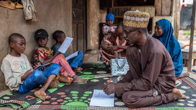 Image of a man and children. The children are reading from exercise books. Image courtesy of the IFPLAN project in Nigeria. The project is UK funded and led by Sightsavers. It focuses on creating equitable access to education, healthcare and employment for people with disabilities.  