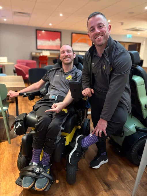 Haydn and Alex are smiling together indoors. Haydn is sitting in his power chair and Alex is crouching beside him. They are both proudly showing their matching purple socks.