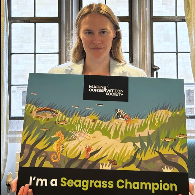 Anna Gelderd MP is stood facing the camera with a window behind her. She's holding a large sign in-front of her which shows a picturesque underwater scene and has the text "I'm a seagrass champion" overlaid. 