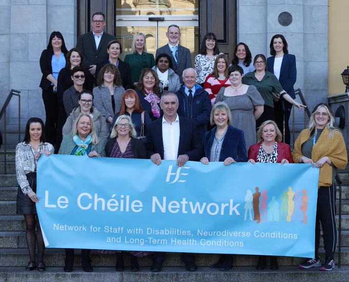 A large group of HSE staff standing on the concrete steps of a grey building. All are smiling at the camera. THe people in the front row are holding a banner that reads: Le Chéile Network. Network for Staff with Disabilities, Neurodiverse Conditions and Long-Term Health Conditions 