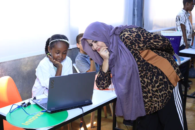 Teacher wearing a purple hijab and leopard print jacket assisting a student working on a laptop in a classroom.