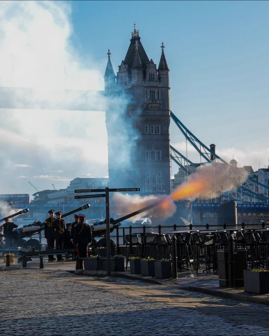 A group of people in ceremonial uniforms participate in a cannon firing ceremony near Tower Bridge in London under a clear blue sky.