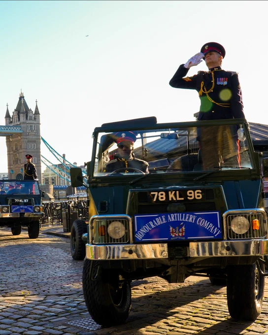 A member of the Honourable Artillery Company salutes from a military vehicle during a parade with the Tower Bridge in the background.