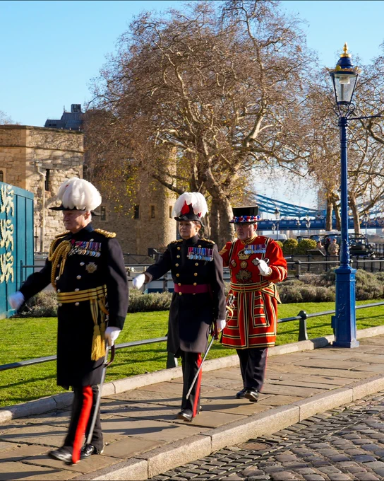 Three members of the British ceremonial guard, including a Beefeater, walking beside the Tower of London on a sunny day.