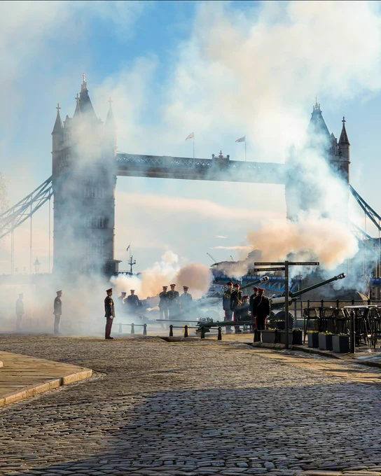 Smoke rises near Tower Bridge as people gather around an area with apparent historical reenactment activities.