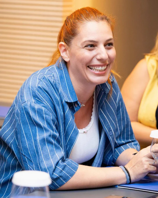 A woman with red hair wearing a blue striped shirt smiles warmly while participating in a classroom-style session.