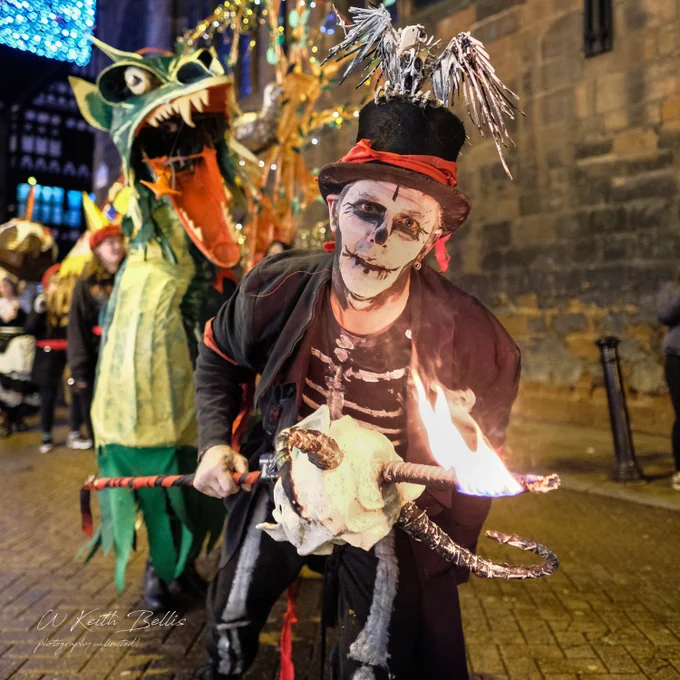 A man dressed as a skeleton holds a flaming torch and looks into the camera. A lifesize dragon puppet roars in the background behind.