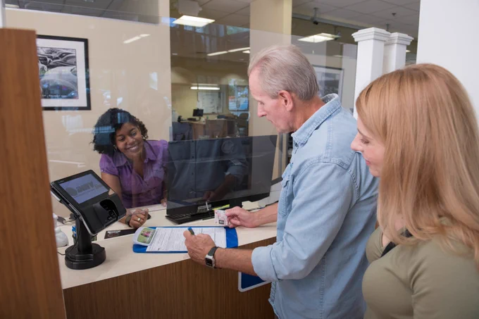 Two people standing at a DMV counter, one of them is signing a form, with a person behind the counter smiling.