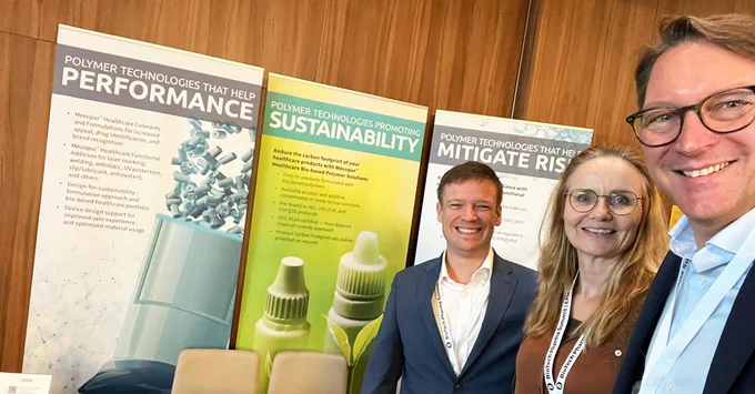 Three smiling individuals, two men and one woman, pose for a selfie at a conference. A man in a blue suit and glasses holds the camera. Behind them are vertical banners for "Polymer Technologies," highlighting performance, sustainability, and risk mitigation.