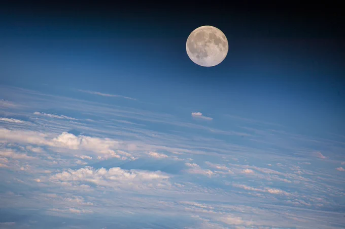 A full moon hangs above the blue glow of Earth's atmosphere as white, fluffy clouds spread across the planet. Credit: NASA