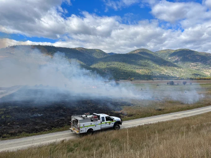 A fire response truck drives along a dirt road beside a smoky field with green mountains under a partly cloudy sky.