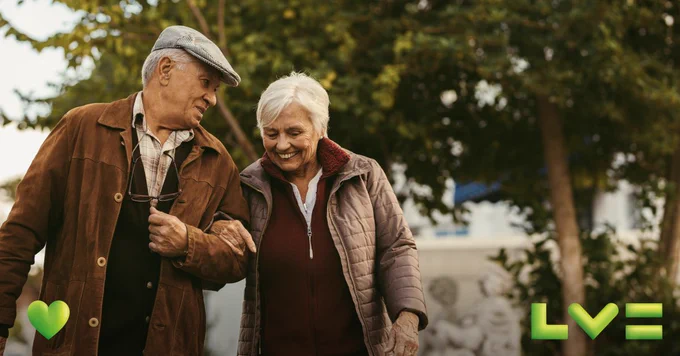 Two older people are walking and smiling together in a park.