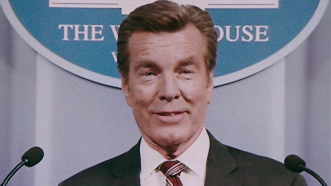 A close up of Peter Bergman as Davis Taffler from Pluribus, appearing on television in the White House press room.