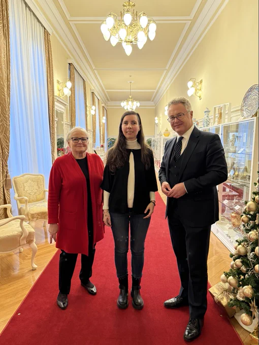 Chargé D’Affaires Nigel Baker, Youth Disability Advocate Mia, and President’s counsellor posing for a photo in the Presidential Palace. There’s a gold and silver decorated Christmas tree on the right, and a longer corridor in the back.