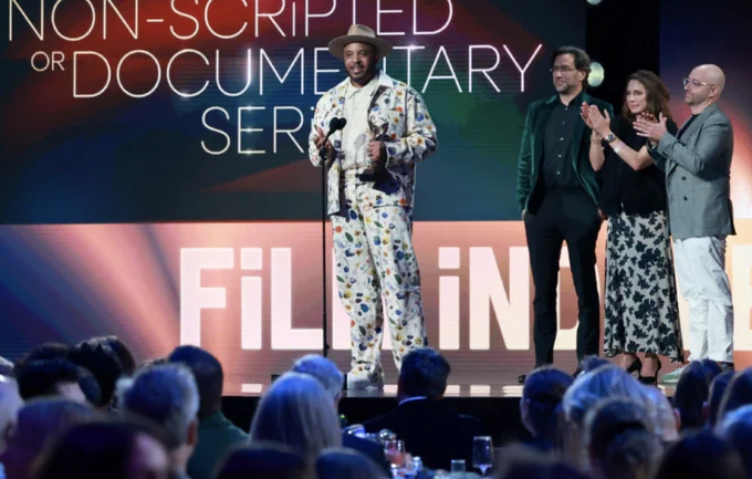 Photo from a past Film Independent Spirit Awards, featuring a man with a hat and off-white suit with colorful designs at a microphone holding an award. Three people are standing behind him on the right, clapping, and in the foreground you can see the top of people in the audience's heads as they watch.
