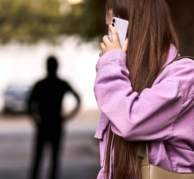 A woman on the street looks behind her at a dark figure stalking her, whilst she holds a mobile phone to her ear