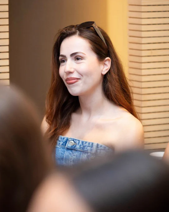 A woman with long brown hair and wearing a strapless denim top smiles softly while attending an indoor event.