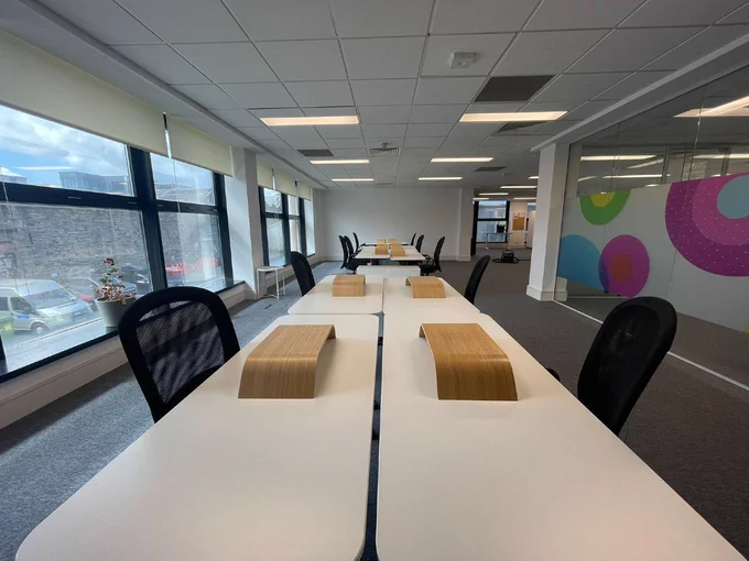 Modern office space with rows of white desks, black chairs and wooden desk organisers. Large windows and a colourful glass partition.