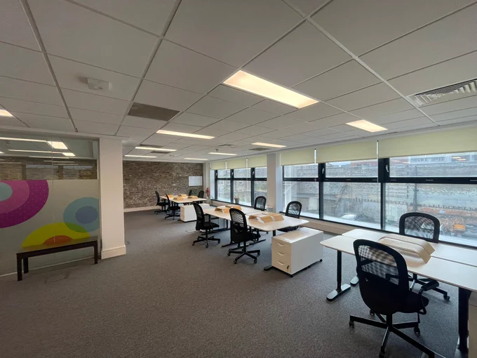 Spacious modern office with multiple desks and chairs, large windows providing natural light and a colourful decorative wall panel.