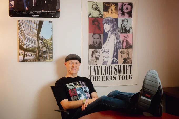 TCU professor of communication studies Andrew Ledbetter sitting on a chair, smiling at the camera, in a room with a large "Taylor Swift: The Eras Tour" poster on the wall.