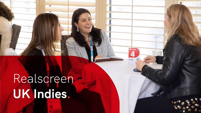 Three women sit at a white-clothed table having a business meeting, looking energised and engaged. In the lower left is a curved, semi-opaque red overlay with the words 'Realscreen UK Indies' in white.