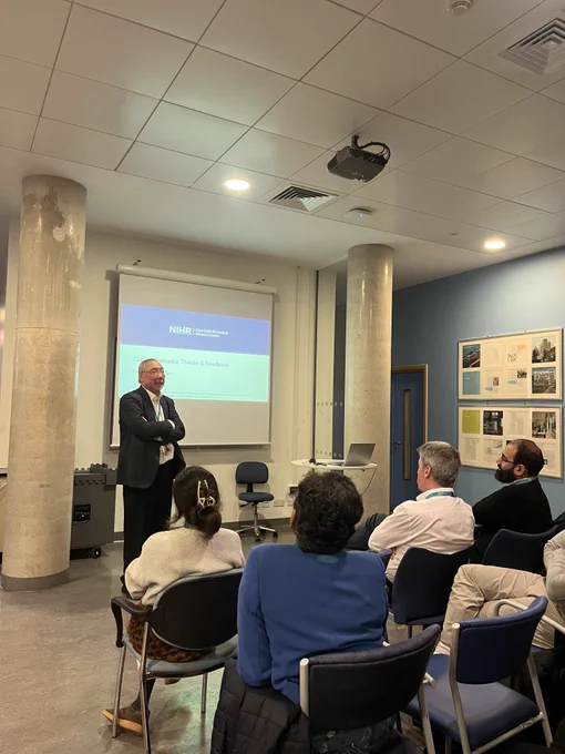 Prof Sir Peng Tee Khaw stood in front of an audience of clinical academics and trainees giving closing remarks at the Ophthalmology Clinical Academic Trainee Forum. Sir Peng is stood in front of a projector displaying the NIHR Moorfields Biomedical Research Centre logo.
