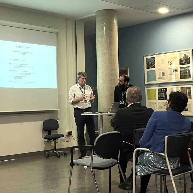 Prof Andrew Webster and Prof Omar Mahroo stood in front of an audience of clinical academics and trainees. Andrew is delivering a talk to the audience. Behind him is a projected agenda for the sixth Ophthalmology Clinical Academic Trainee Forum.