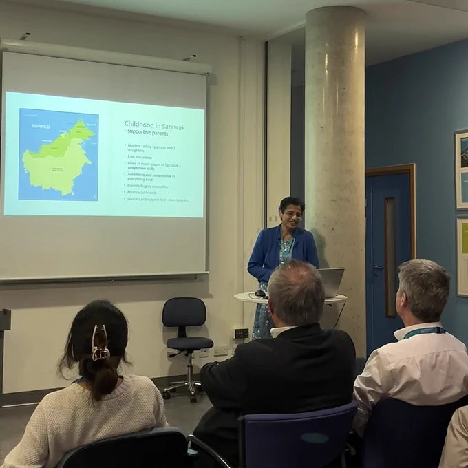 Prof Sobha Sivaprasad stood in front of an audience of clinical academics and trainees delivering a talk on her clinical academic journey. Behind her is a projector displaying a slide detailing Sobha's early life in Sarawak, Malaysia.