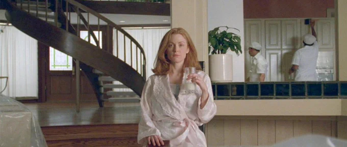 Still from SAFE of a giner woman in a pink dressing gown. She holds up a glass of milk in one hand, and rests the other on a book. She is in a large living room with iteams covered in plastic sheets, and behind her are two men in overalls paining a door.